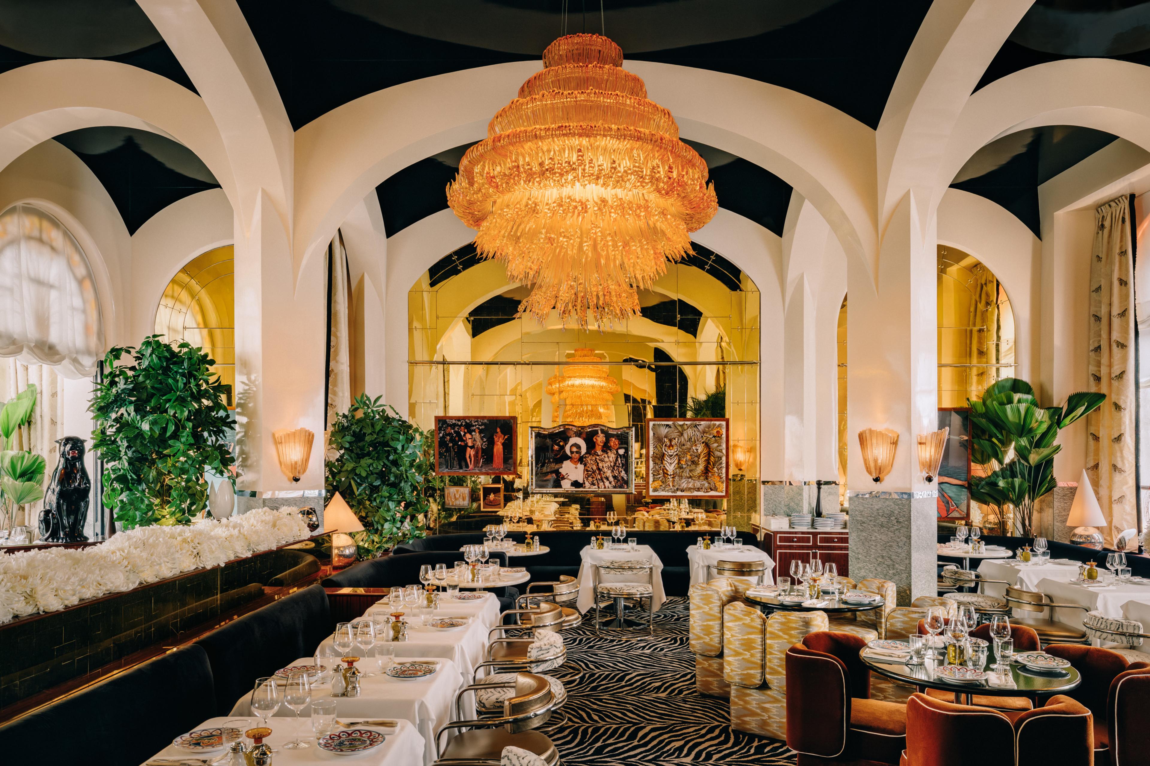 Full dining room of Gloria Paris with vaulted white arches, a large tiered amber chandelier, zebra-print carpet, tropical plants, black banquettes and gold-framed artworks reflected in a mirrored back wall.