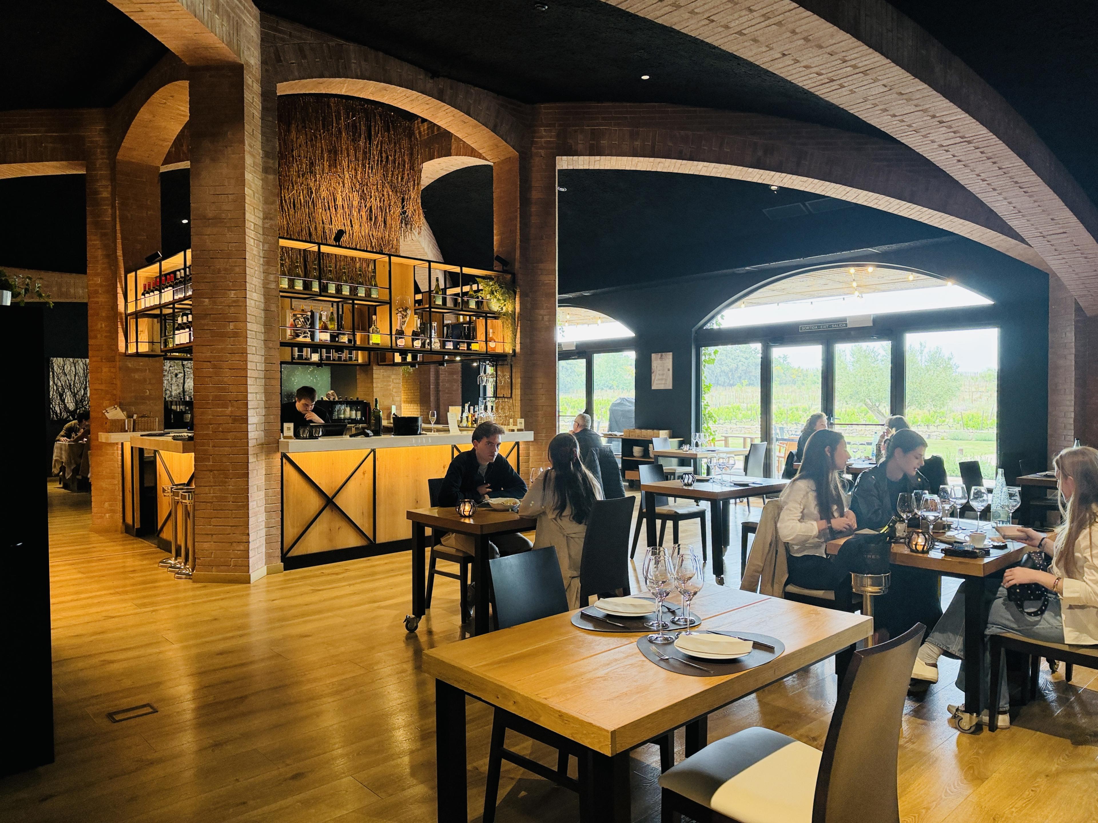 the warm brick-arched dining room at Jardí restaurant in Pinedes, with diners seated at wooden tables and vineyard views through large windows