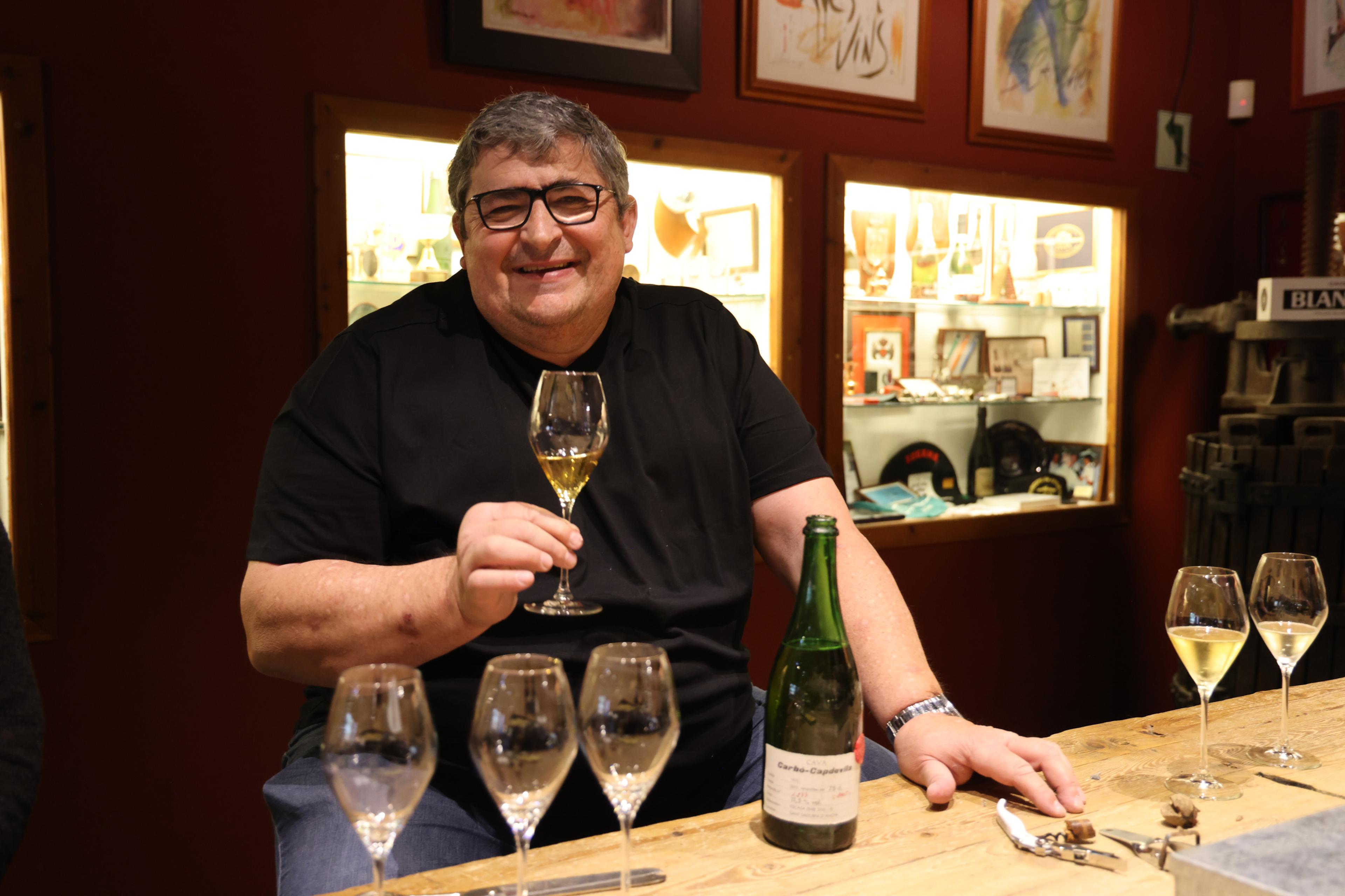 A smiling man holds a glass of aged Cava beside an open bottle of Carbó-Capdevila 1975 during a tasting at Blancher's Pinedes cellar.