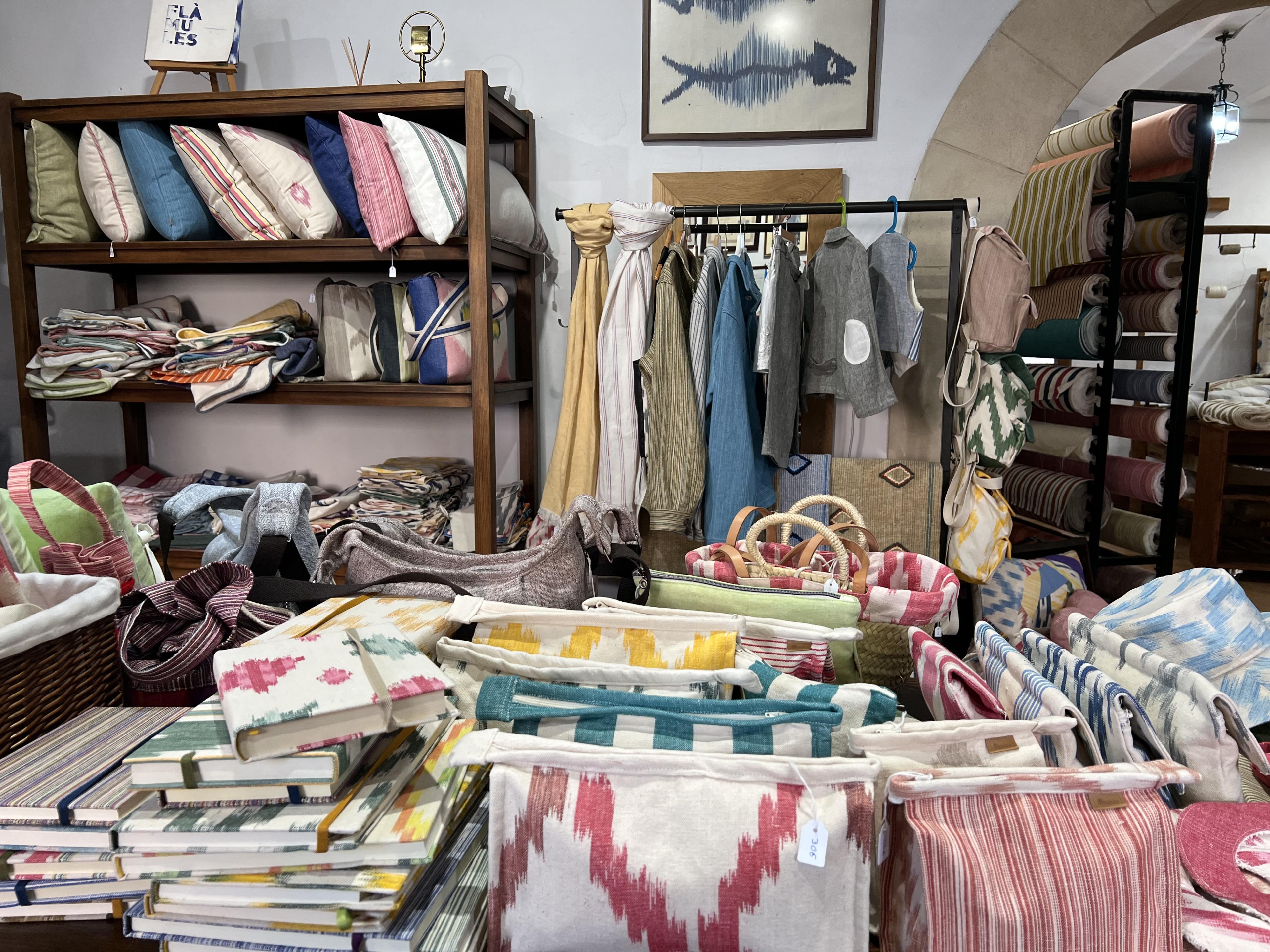 Textile shop with pillows on shelf, scarves and clothing on a rack, and a table with bags and journals